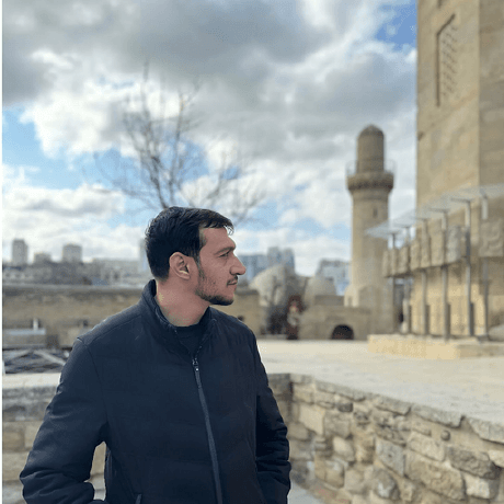 Ali Hassan in a dark winter jacket, looking off to one side, standing in a stone courtyard with a minaret and cloudy sky behind him.